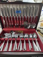 Full view of stainless steel cutlery set arranged inside vintage wooden box with red felt lining, showing knives, forks, spoons, serving utensils.