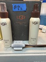 Photo of two UGG spray bottles (Sheepskin Protector and Freshener) and a cleaning brush with brown handle, with UGG branded booklet behind them.