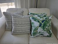 Four accent sofa cushions resting on a light beige upholstered sofa: two beige/brown geometric patterned with removable cases, two with green palm leaf print without removable cases.