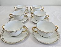 Top angle view of six porcelain soup bowls with two handles each, paired with matching saucers. All feature a floral band with pink and green flowers and gold trim on edges.