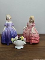 Two Royal Doulton figurines in detailed 19th-century style dresses: one purple, one pink, with a small floral ceramic arrangement in front