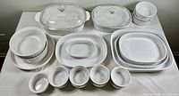 Full view of all white glass Corningware items arranged on table including large rectangular and oval dishes with lids, pie dish, and multiple bowls.