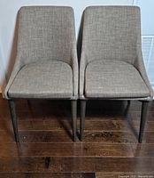Front view of two matching beige fabric dining chairs with black legs showing full front and seat area.