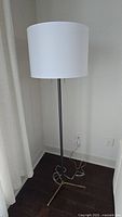 Full view of the floor lamp showing the white drum shade, black metal pole, and brass tripod base.