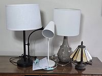 Four table lamps arranged side by side on table against wall, showing full lamps and shades