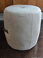 Side view showing the round fabric ottoman with a brown leather handle attached with rivets.