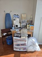 Wide shot showing ironing board, wooden sewing cabinet with Kenmore sewing machine, plastic organizer drawers, compartment box, needlework books, basket and bag of batting.
