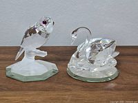 Two Swarovski crystal figurines on a wooden surface against a wall: parrot on branch and swan.