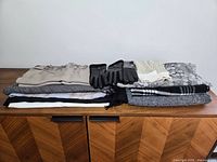 Photo showing an assortment of scarves and gloves neatly folded on a wooden surface. Various neutral colors and patterns appear with some fringed edges visible.