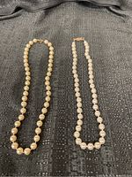 Two strands of pearl beads laid flat on a black textured surface. One strand is cream-colored, the other is white.