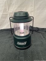 Front view of dark green Coleman camping lantern with clear plastic globe and handle, placed on a black surface with white background.