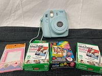Light blue Fujifilm Instax Mini 8 instant camera with wrist strap, three boxes of Fujifilm Instax Mini instant film, and one pink mini photo frame arranged on a black surface.