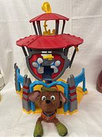 Paw Patrol Lookout Tower with red roof, bone emblem, blue and gray structure, yellow slide and ladder, with plush Chase dog in front.