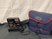 Polaroid Autofocus 660 instant camera with lens, flash, and strap visible, alongside blue and red fabric carrying case.