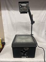 Black EIKI still picture projector with extended adjustable arm and lens housing, showing flat glass projection surface and unit base with power cable.