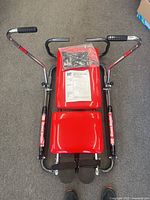 Top-down view of the red DP Shape Master 1000 rowing machine showing its padded seat, backrest, foot straps, and dual curved handlebars with grips on a grey carpeted floor.