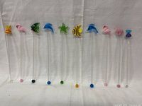 Set of 10 vintage glass swizzle sticks arranged in a row showing various animal and candy glass figures on top of each clear rod.