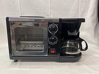 Full front view of multifunction breakfast machine showing toaster oven with glass door on left, control dials in middle, power switch, and attached coffee maker with glass carafe on right.