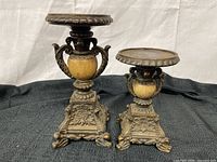 Two vintage candle holders on black fabric background with beige urn shapes and ornate metal bases.