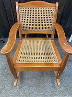 Front view of the vintage wooden child's rocking chair showing cane seat and back with armrests and rockers.