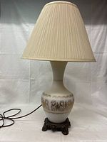 Front view of the vintage ceramic table lamp with a beige pleated fabric lampshade and ornate dark metal base. Electrical cord visible.