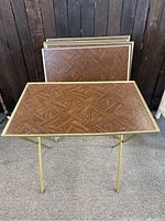 Set of six vintage TV trays stacked and displayed, showing wooden patterned tops and gold-tone legs.