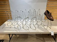 Nineteen wire shoe display stands arranged on a table against a wall, all empty except one holding a brown leather shoe.