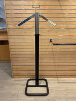 Full view of the 2-way metal clothing rack against wooden slatted wall, showing the black metal base and structure with dual arms equipped with hooks.