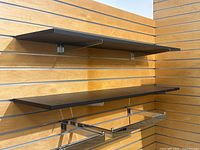 Two black wall-mounted shelves displayed on wooden slatwall panels. Shelves supported by sturdy silver metal brackets. Visible minor wear on edges and surface. Shelves measure approximately 48 inches wide, 12 inches deep, and 3 inches high.