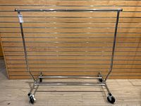 Full view of collapsible clothing rack in front of wood slat wall. Chrome metal with four wheels and extending arms visible.