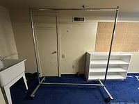 Full front view of the extra tall metal clothing rack standing against a wall next to a white shelving unit and utility sink.