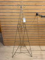 Front view of folding gold metal artist easel showing full triangular metal frame and fleur-de-lis top design against wood slat wall.