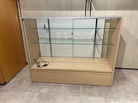 Front view of glass display case showing wood base, two glass shelves and sliding glass doors with lock, electrical cord on bottom shelf.