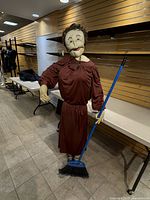 Full body image of the life-sized doll standing in a store environment holding a blue broom, wearing a burgundy dress and wooden necklace.
