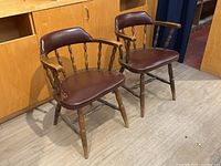 Front angle photo showing pair of vintage pub style spindle back armchairs with burgundy faux leather padded seats and backs, worn upholstery and wood finish.