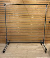 Front view of the heavy duty metal rolling clothing rack against a wooden wall backdrop on a wood floor.