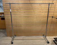 Full frontal view of the metal rolling clothing rack against a wooden slat wall background on caster wheels.