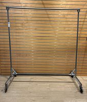 Full front view of the heavy duty metal clothing rack showing its rectangular base, vertical supports, and horizontal hanging bar against a wooden slat wall.