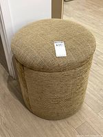 Full view of the round foot stool ottoman showing beige woven fabric upholstery and cylindrical shape.