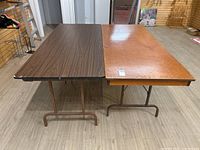 View of two folding tables side-by-side showing wood veneer tops and tubular metal legs.