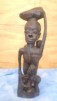 Full view of the dark wooden statue showing the mother holding a bowl on her head and a child standing at the base.