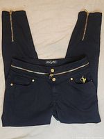Front view of black Baby Phat jeans showing gold zipper at waist, gold button closure, and embroidered logo on pocket.