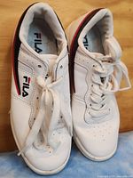 Pair of white Fila women's sneakers with black and red stripes shown from a top view on a wooden surface.