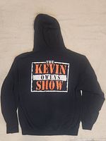 Back view of black WWE Kevin Owens hoodie showing a large rectangular graphic saying 'THE KEVIN OWENS SHOW' in distressed orange and white print on the back.