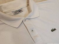 Close-up of upper front with collar, buttons, and embroidered green crocodile logo on white cotton fabric