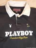 Close-up of collar, button placket, and front logos including Playboy bunny, OVO owl, large white PLAYBOY text and gold October's Very Own script.