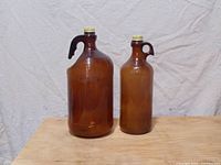 Front view of two vintage dark amber glass bleach bottles with metal screw caps and handles, one larger with ridges and one smaller.