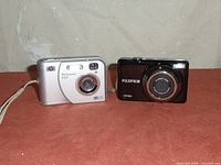 Front view of two digital cameras, HP Photosmart E237 in silver on left and black Fujifilm digital camera on right.