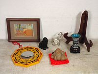 Overview of lot items arranged on flat surface including Feng Shui mirror, Buddha figurine, dog figurine, incense holder, decorative vase, wooden plate holder, framed painting.