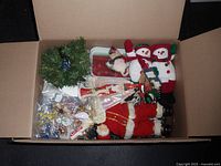 Open cardboard box containing assorted Christmas decorations including plush snowman and Santa figurines, small wreath, ornaments, and other items